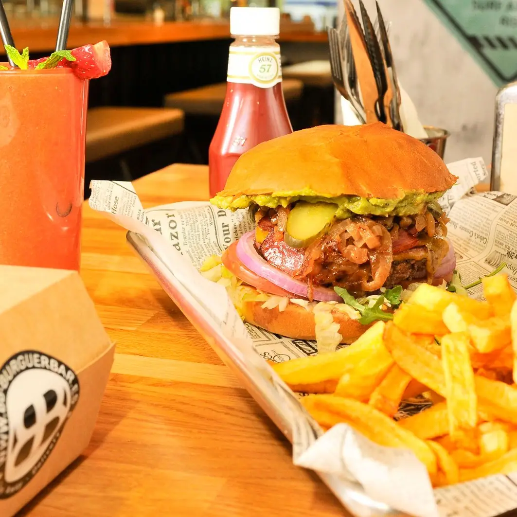 Hamburguesa con cebolla crujiente, guacamole y pepinillos sobre tabla de madera, con patatas fritas y un batido de zanahoria
