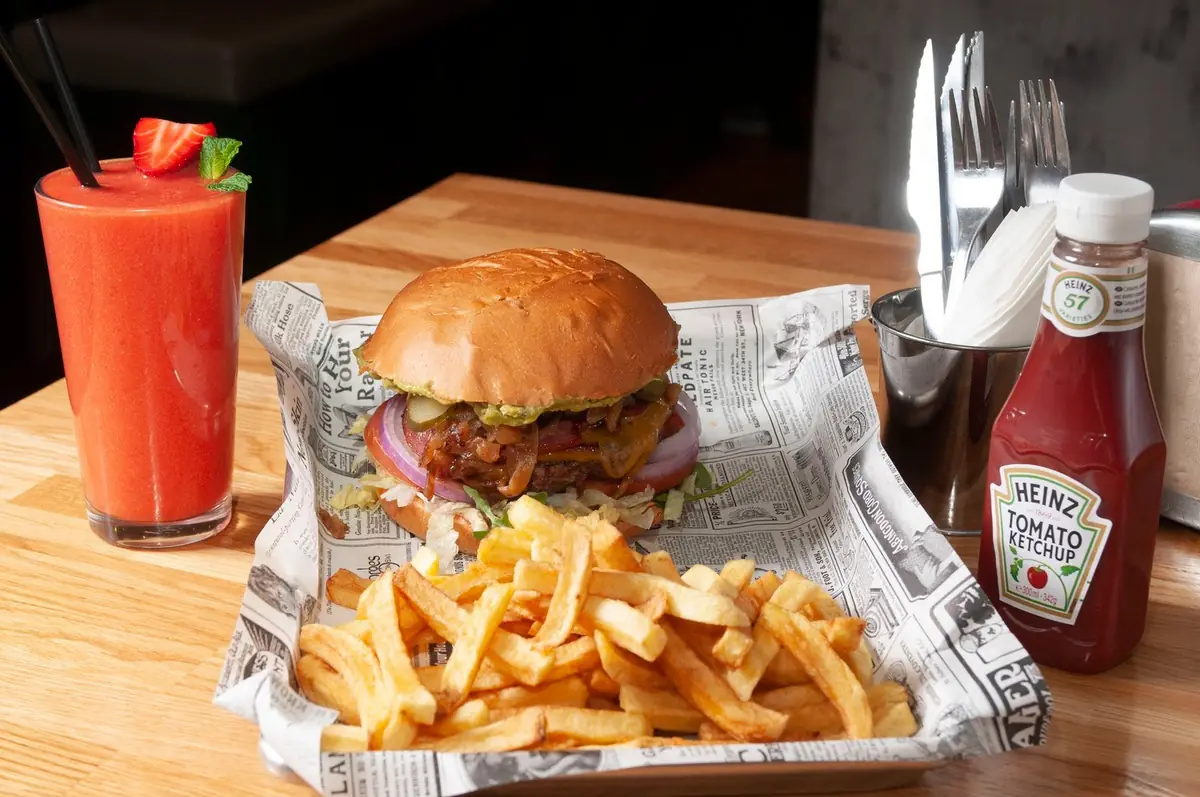 Hamburguesa con bacon y cebolla morada servida sobre papel de periódico, con patatas fritas y un batido de fresa