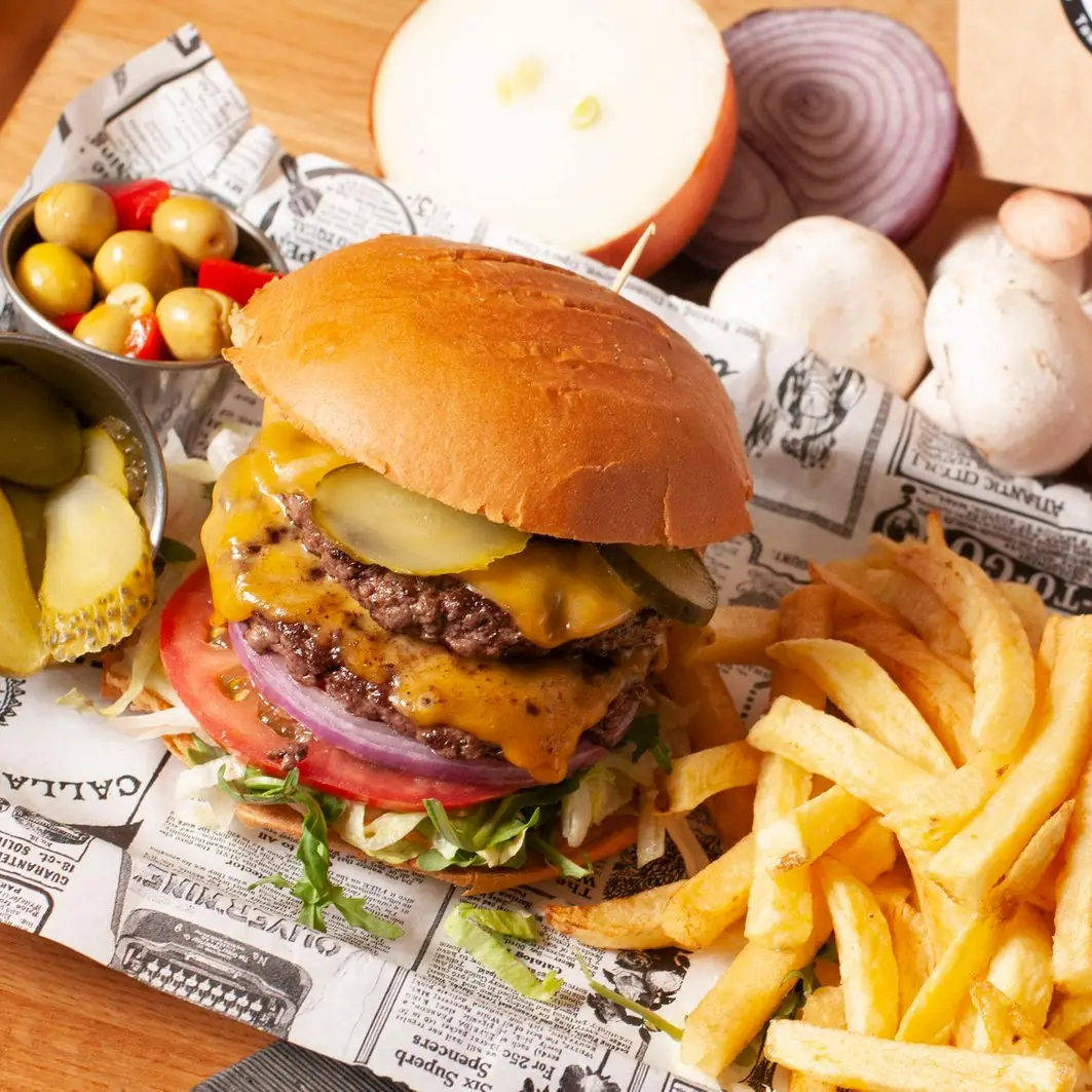 Hamburguesa doble de Angus con cheddar fundido, pepinillos y tomate, acompañada de patatas fritas y un cuenco de aceitunas