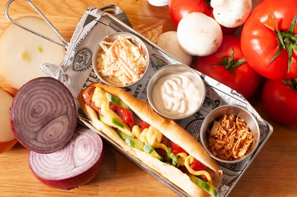 Perrito caliente con mostaza, pepinillo y pimiento en bandeja metálica, con coleslaw, salsa blanca y cebolla frita para mojar