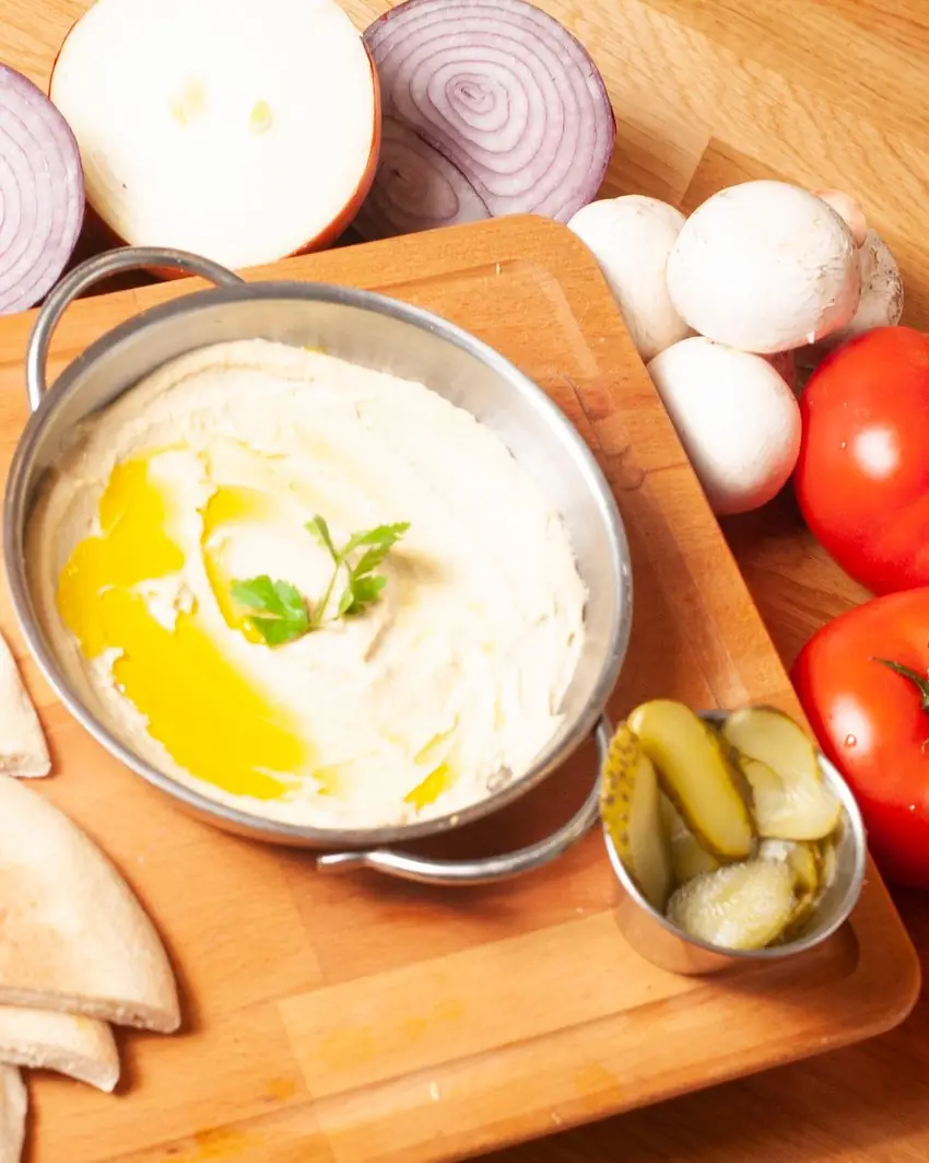 Hummus con un hilo de aceite de oliva, servido en tabla de madera con pan de pita y pepinillos