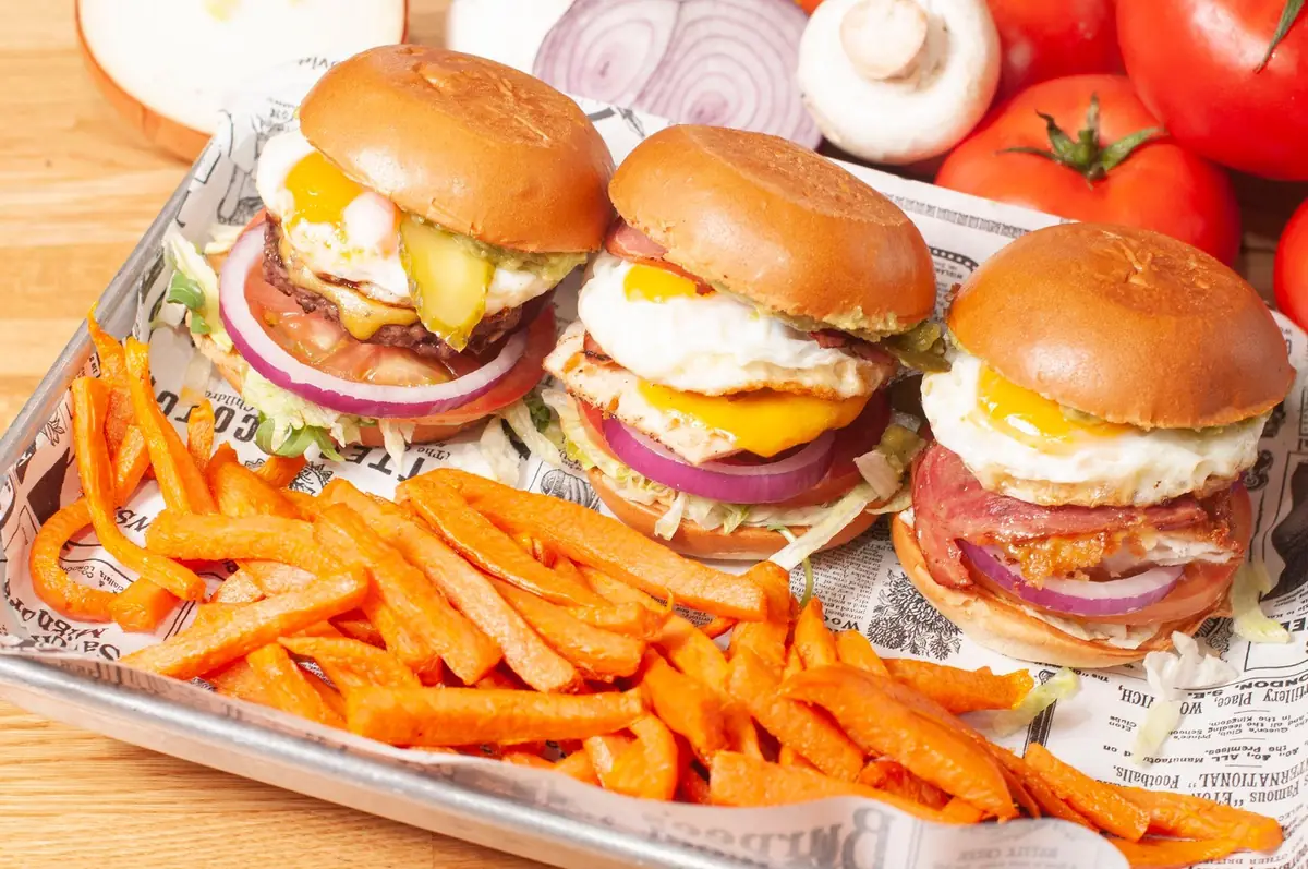 Tres mini hamburguesas coronadas con huevo frito y bacon, junto a una montaña de patatas de boniato